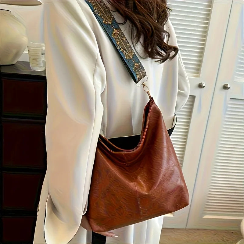 A woman holds a brown leather shoulder bag with a wide blue embroidered strap, worn under a white blazer.