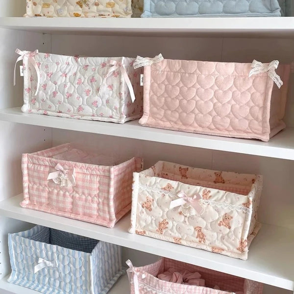 A display of pink and white quilted cotton storage baskets with lace flower designs on white shelving.