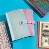 Une pile de trois carnets scintillants roses et bleus à côté d'un ordinateur portable et d'une pile de monnaie américaine.