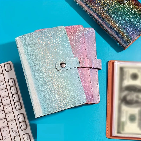 A stack of three glittery pink and blue notebooks next to a laptop and a stack of US currency.