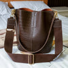 A brown alligator pattern leather crossbody bag resting on white bedding.