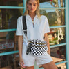 A woman wearing a white polo shirt and shorts stands in front of a glass door, holding a checkered crossbody sling bag.