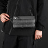 A person wearing a black jacket holds a black versatile fashion chest bag with a strap.