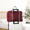 Sac de voyage pliable marron avec poignées noires posé sur une valise à roulettes blanche.