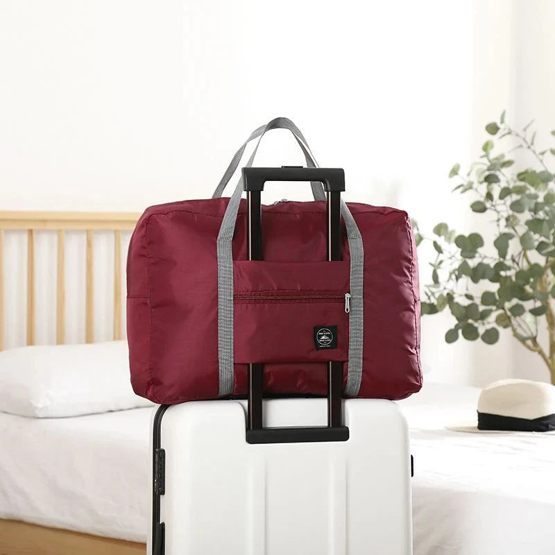 Sac de voyage pliable marron avec poignées noires posé sur une valise à roulettes blanche.