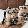 A fluffy brown bear plush toy accessorized with a camera and black glasses, resting on a brown surface.