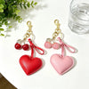 A pair of pink and red heart-shaped keychains with gold clasps and cherry pendants resting on a white surface.