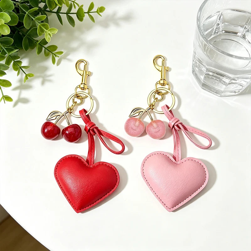 A pair of pink and red heart-shaped keychains with gold clasps and cherry pendants resting on a white surface.