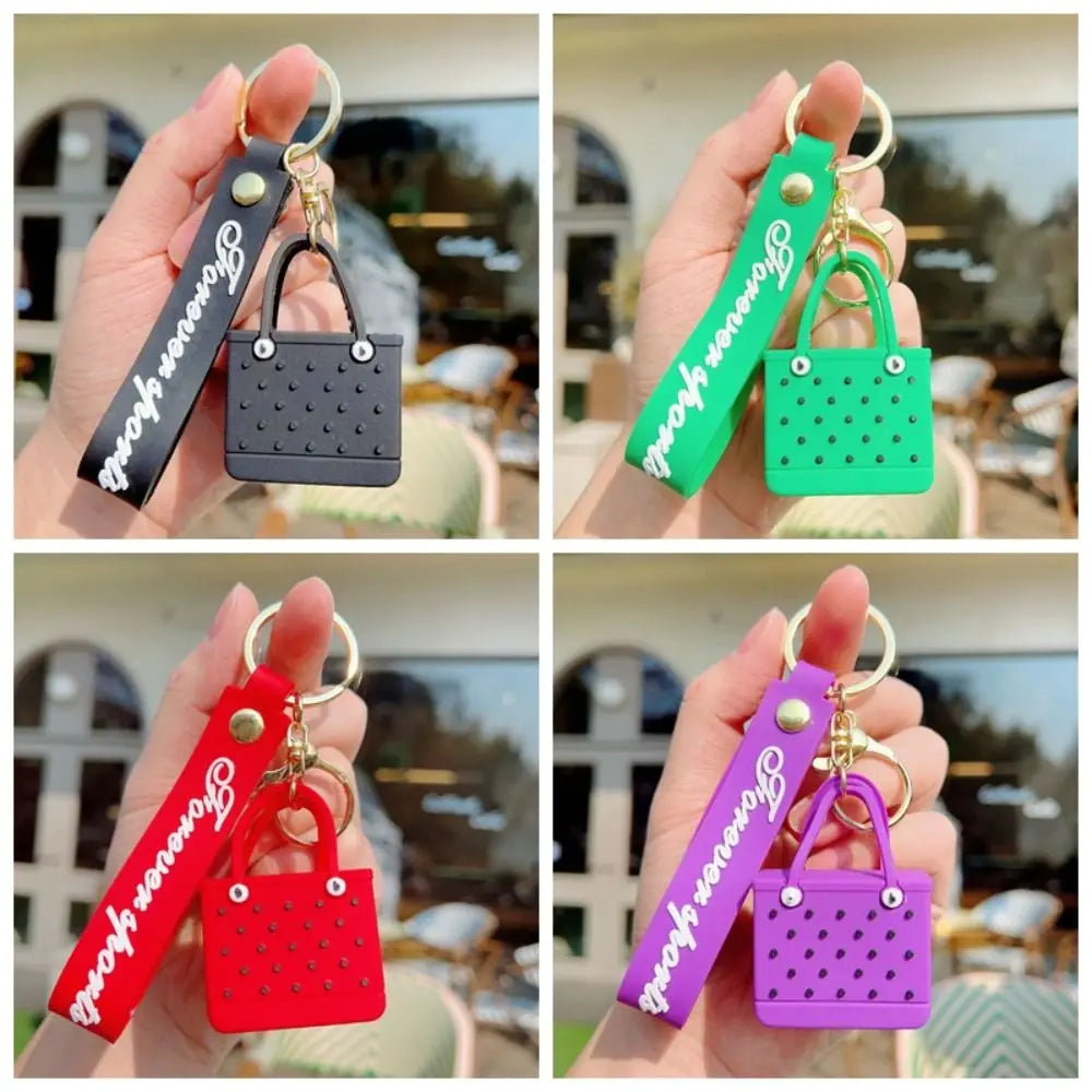A hand holds a silicone heart beach bag keychain in various colors including black, green, red, and purple.
