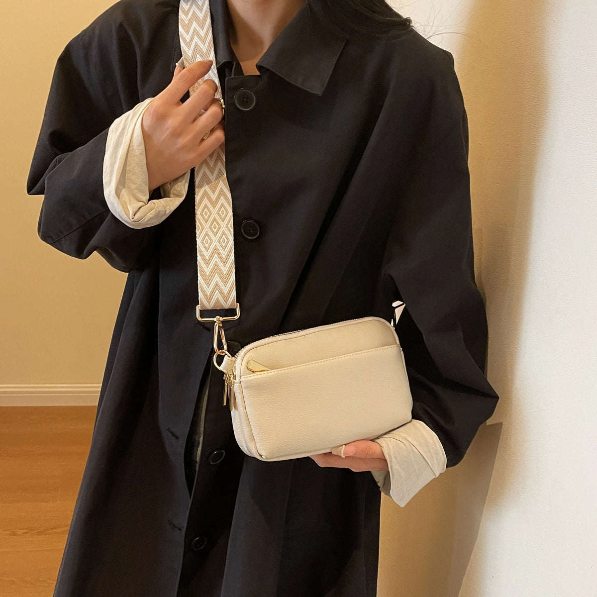 A woman wearing a black coat holds a cream vegan PU crossbody bag with a patterned strap.
