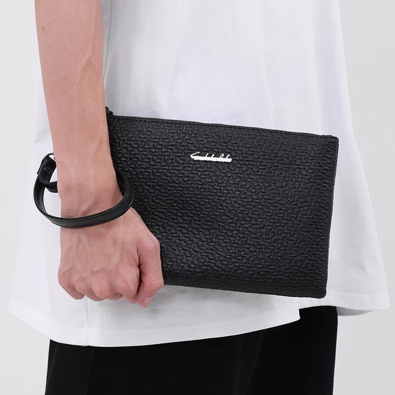 Eine schwarze Kunstleder-Herrenclutch wird von einer Person in einem weißen Hemd gehalten.