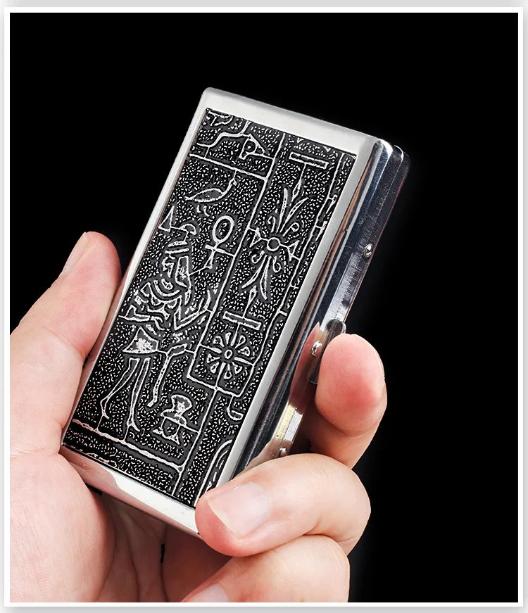 A hand holds a silver metal cigarette case holder featuring intricate black engraving.