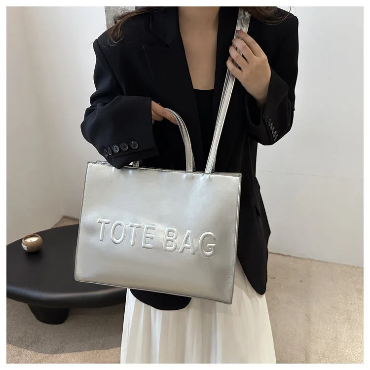 A woman holds a silver tote bag with a leather strap against a white background.