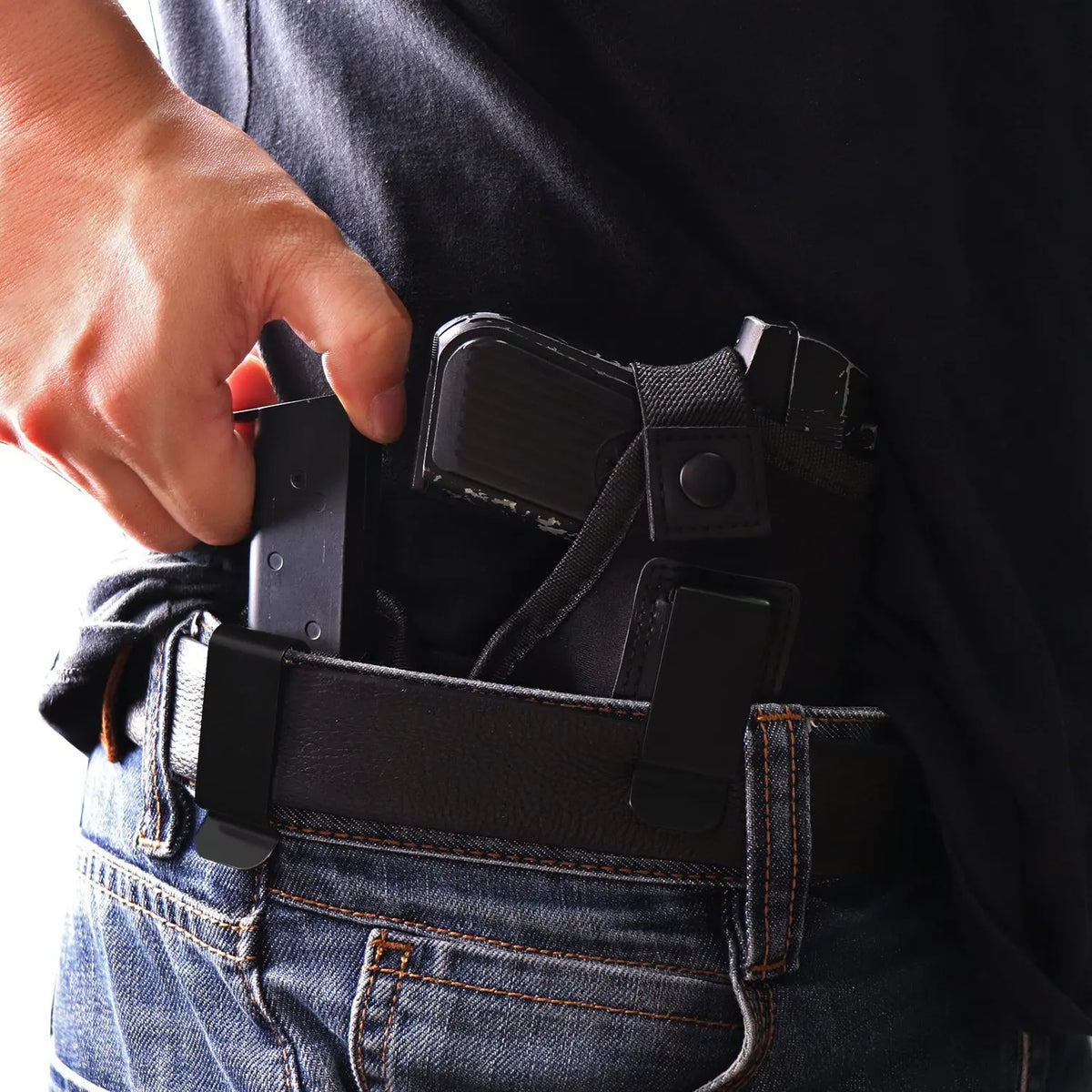A hand is inserting a black universal inside-the-waistband magazine holster into the waistband of blue jeans.