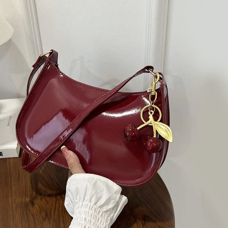 A deep burgundy PU leather shoulder bag with a gold cherry pendant is held by a hand against a white background.