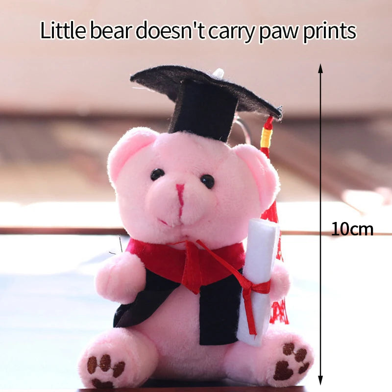 Pink plush bear keychain wearing a graduation cap, red ribbon, and holding a white object.
