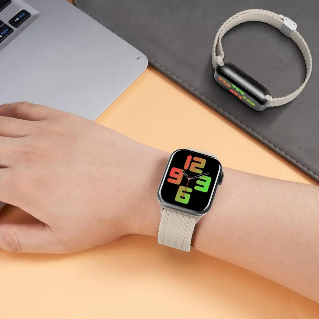 A wrist wearing a black smartwatch with a light beige braided stretch band resting on a gray surface.