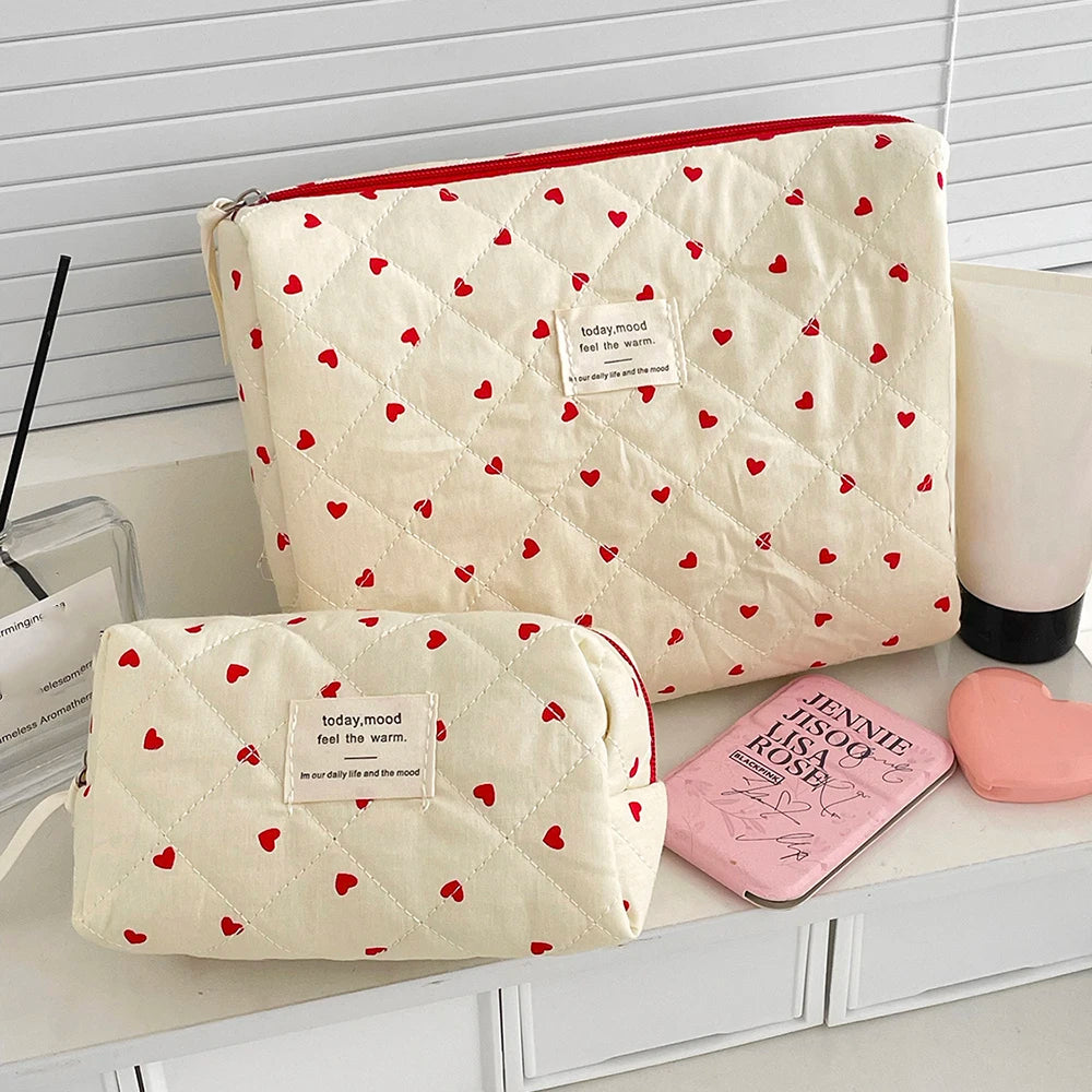A cream quilted makeup bag with red hearts and a red trim, next to a pink compact.