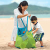 A woman is holding a green mesh storage bag for toys while kneeling on the beach with a young boy.