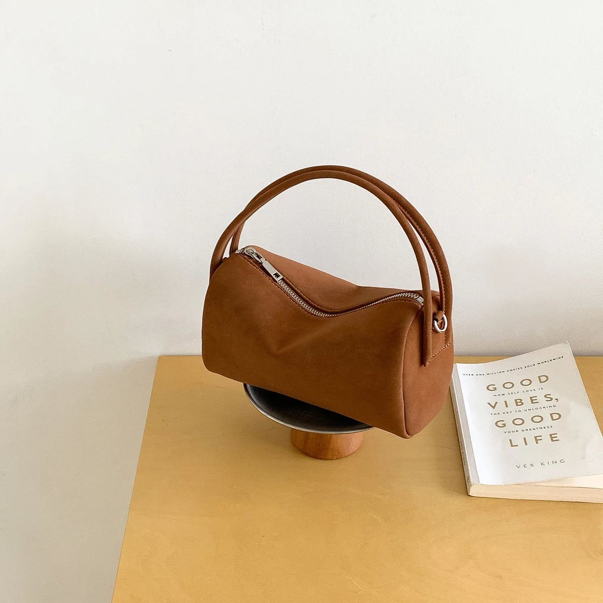 A tan faux suede crossbody bag resting on a light wood block against a white background.