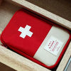 A red compact survival and first aid kit with a white cross on a light background inside a wooden box.