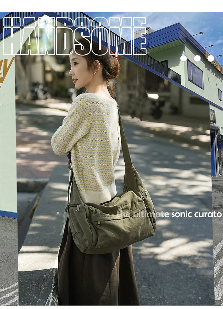 A woman wearing a cream knit sweater and dark skirt holds a large olive green cotton canvas shoulder crossbody bag.