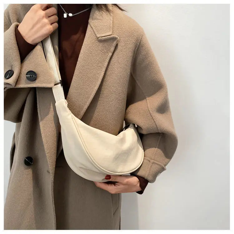 A woman wearing a beige wool coat is holding a cream-colored preppy sling crossbody bag.
