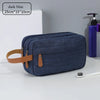 A dark blue portable travel storage pouch with a tan strap is shown on a white surface next to a toothbrush and a purple bott