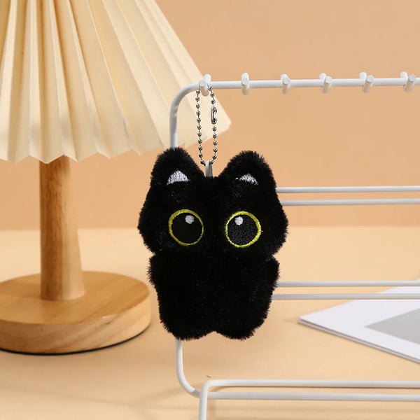 Un charm de chat en peluche noir pend d'un porte-clés en fil d'argent sur un présentoir métallique blanc.
