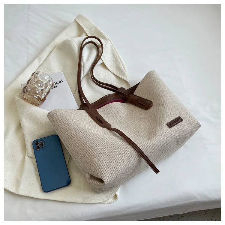 A beige canvas shoulder tote bag with brown leather straps and a smartphone resting on the fabric.