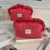 A red and white checkered makeup pouch with a lace trim is displayed on a white surface next to books and makeup brushes.
