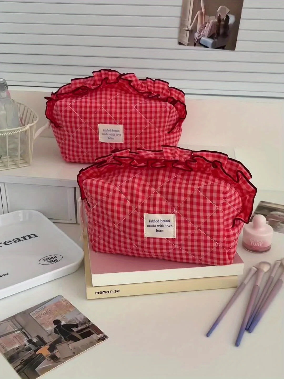 A red and white checkered makeup pouch with a lace trim is displayed on a white surface next to books and makeup brushes.