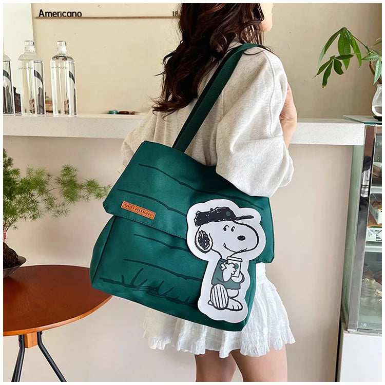 A woman holds a dark green canvas tote bag featuring a white Snoopy design in a casual indoor setting.