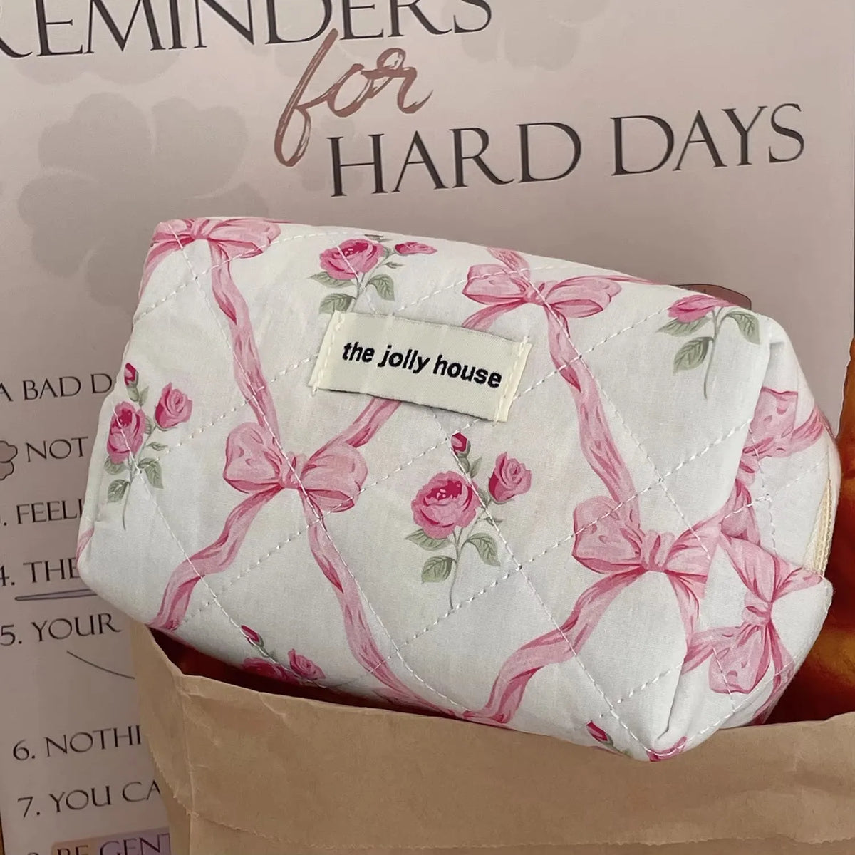 A quilted pink and white floral embroidered makeup bag with a bow, displayed on a brown paper background.