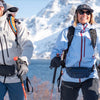 Two women hiking in snowy mountains with backpacks and a water view.