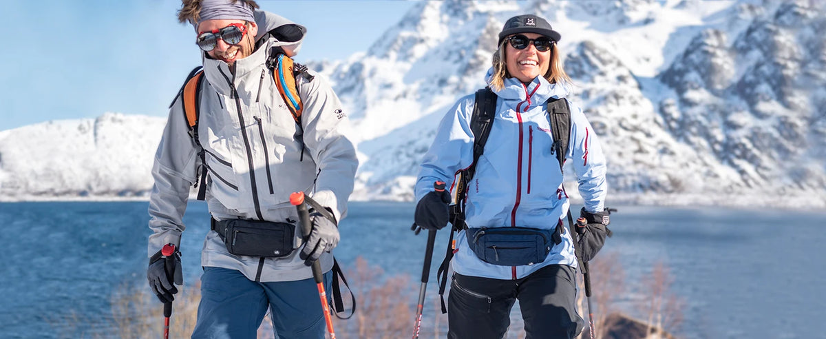 Two women hiking in snowy mountains with backpacks and a water view.
