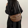 A brown woven pattern waist bag with a gold chain strap is held by a person wearing a black coat.