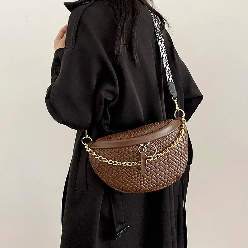 A brown woven pattern waist bag with a gold chain strap is held by a person wearing a black coat.