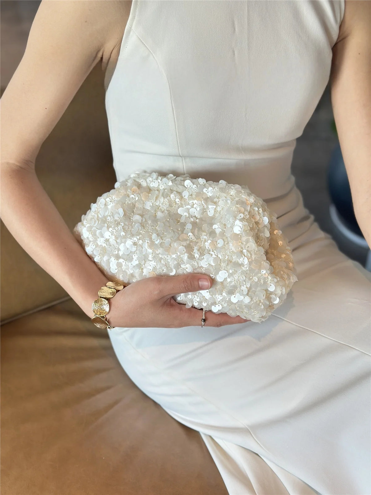 A woman holds a white beaded chain shoulder clutch bag against a white dress.