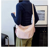 A woman wearing a dark blue top and olive pants holds a pink canvas crossbody bag against a white wall.
