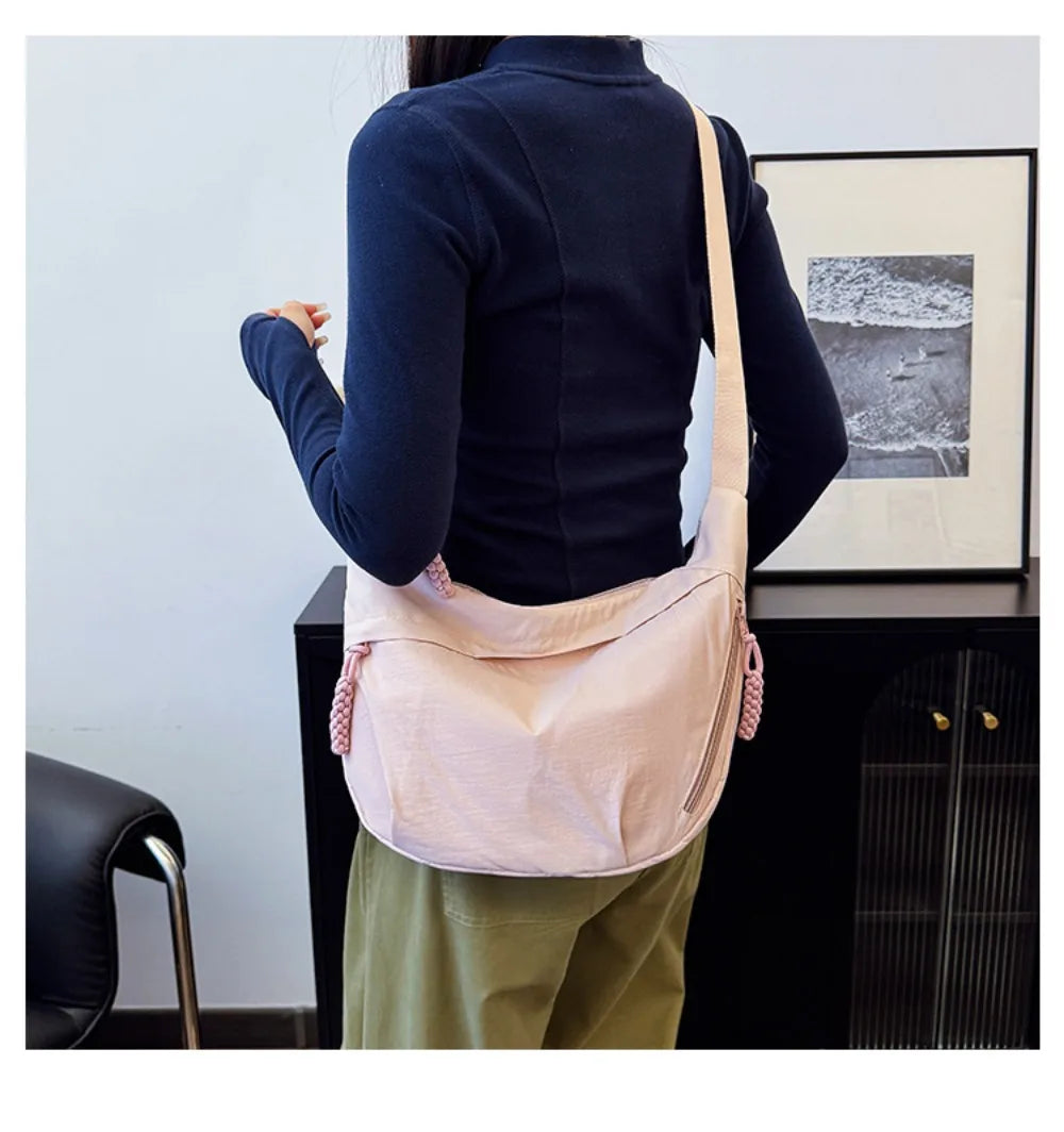 A woman wearing a dark blue top and olive pants holds a pink canvas crossbody bag against a white wall.