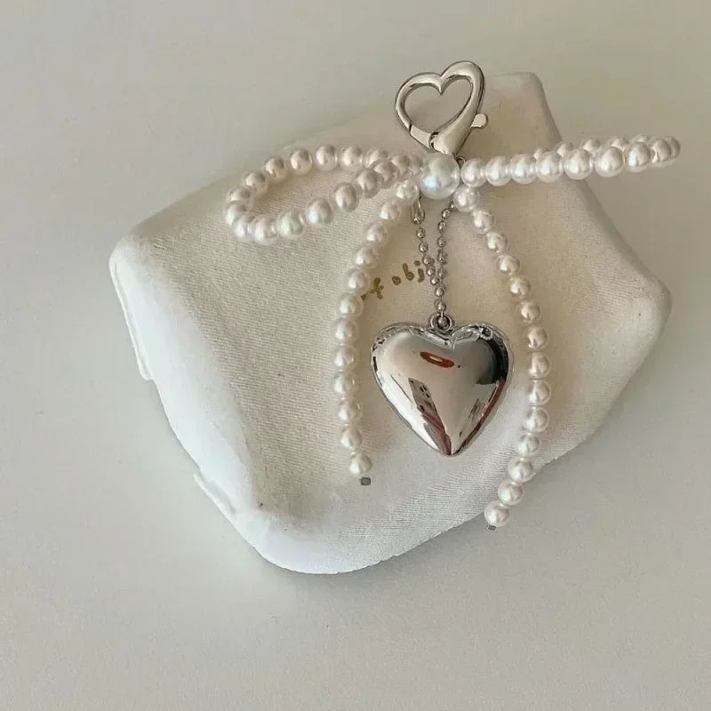 A silver heart charm suspended from a pearl beaded bowknot keychain displayed on a white surface.