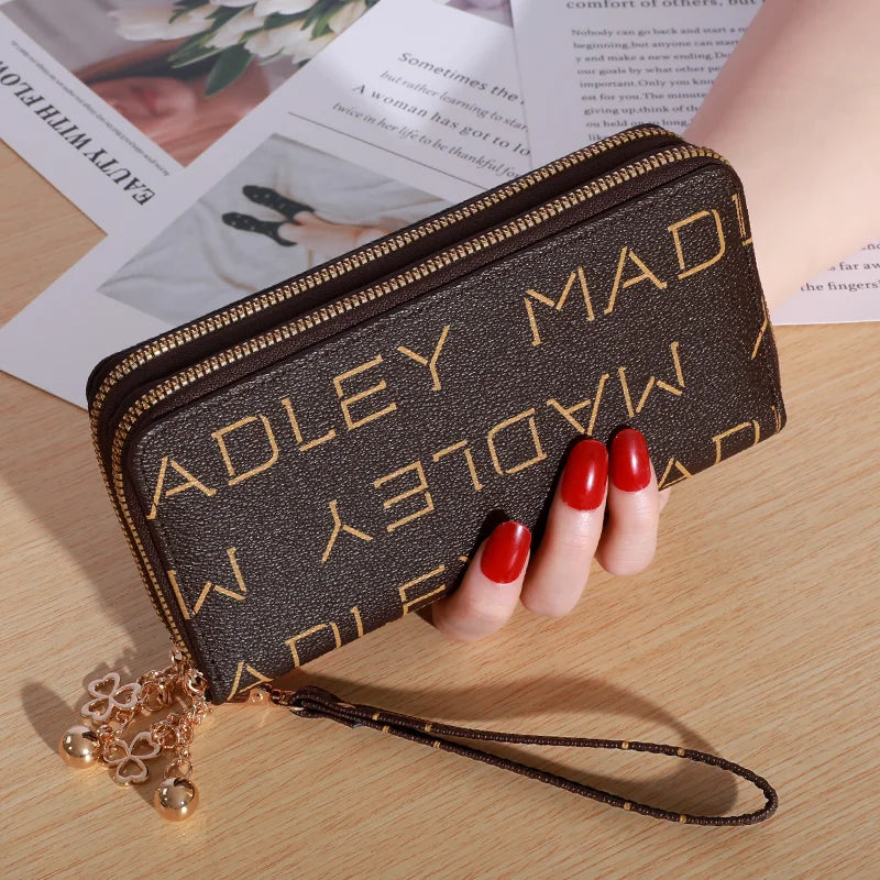 Cartera con cremallera de cuero sintético marrón oscuro y costura dorada, con correa de cuero, sobre una superficie de madera