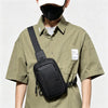 A person wearing a black face mask and a olive green shirt holds a black chest bag.