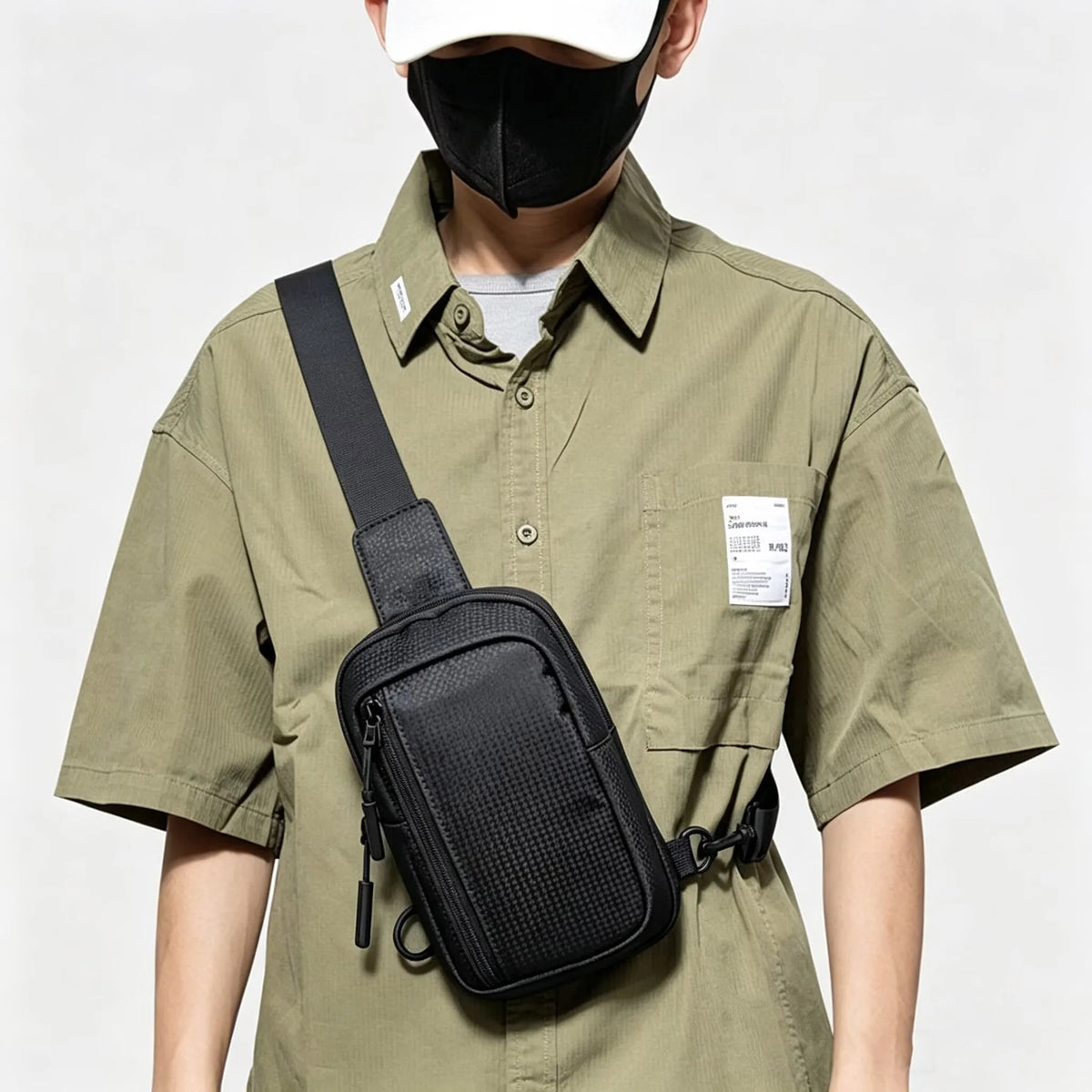 A person wearing a black face mask and a olive green shirt holds a black chest bag.