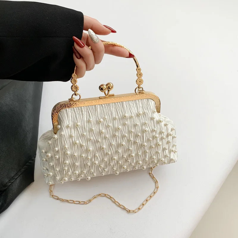 A white pearl embellished crossbody shoulder bag with gold hardware being held by a person's hand.
