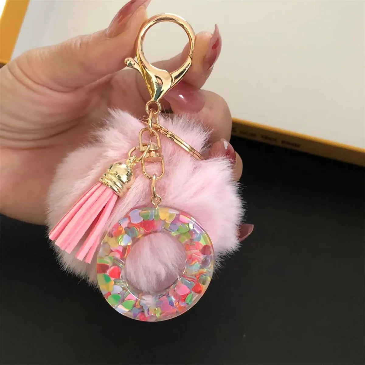 A gold-colored keychain featuring a pink faux fur pompom and a clear resin ring with colorful confetti.