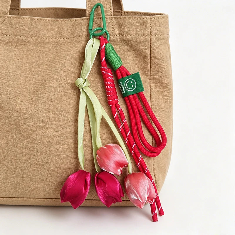 A beige tote bag with a green and red tulip flower charm keychain hanging from the strap.