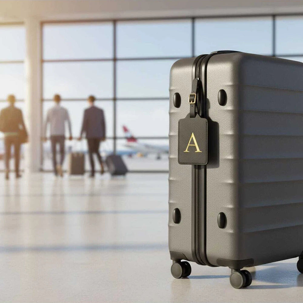 A gray PU leather suitcase with a gold letter A luggage tag on the front in an airport-like setting.