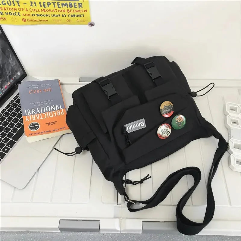 A black nylon crossbody shoulder bag is displayed on a white desk next to a laptop and a book.